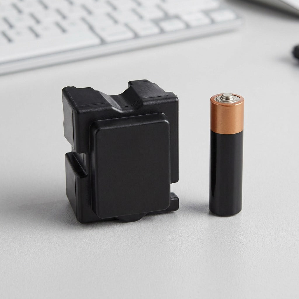 A single black Xerox ColorQube ink stick is pictured next to a standard AA battery on a light grey, slightly textured desk surface under clean, even studio lighting. A person's hand is visible just to the side, further contextualizing the scale. This comparison provides a direct visual reference to the actual size of the solid ink stick.